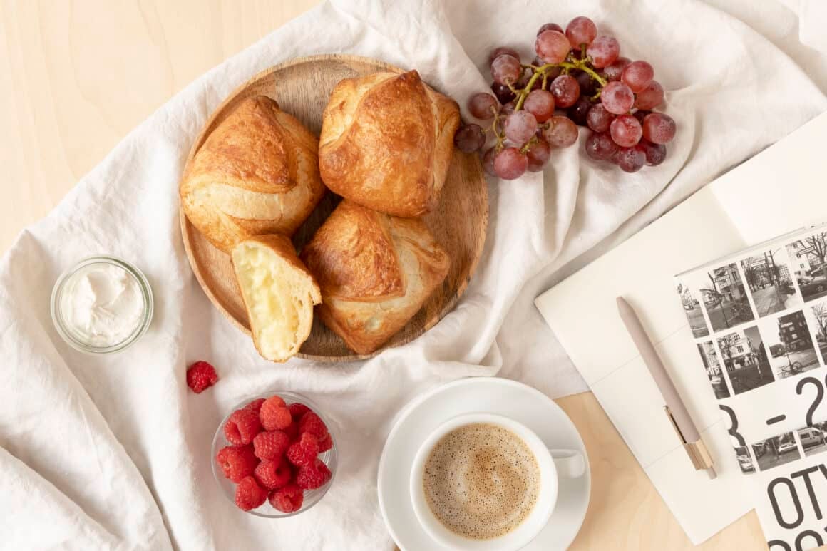 Auf einem hellen Tuch steht ein Holzteller mit veganen Plundertaschen. Daneben liegen Trauben, eine Schale mit Himbeeren, ein Frischkäsedip und eine Tasse mit Kaffee