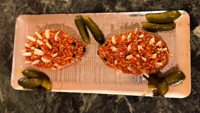 Zwei vegane Mett Igel liegen auf einer Platte. Dekoriert mit Zwiebeln als Igel Stacheln und Essiggurken liegen darum verteilt
