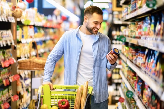 Mann steht in einem Gang eines Supermarktes. Er schiebt einen Waagen vor sich und liest sich den Text auf einer Tomatensauce durch
