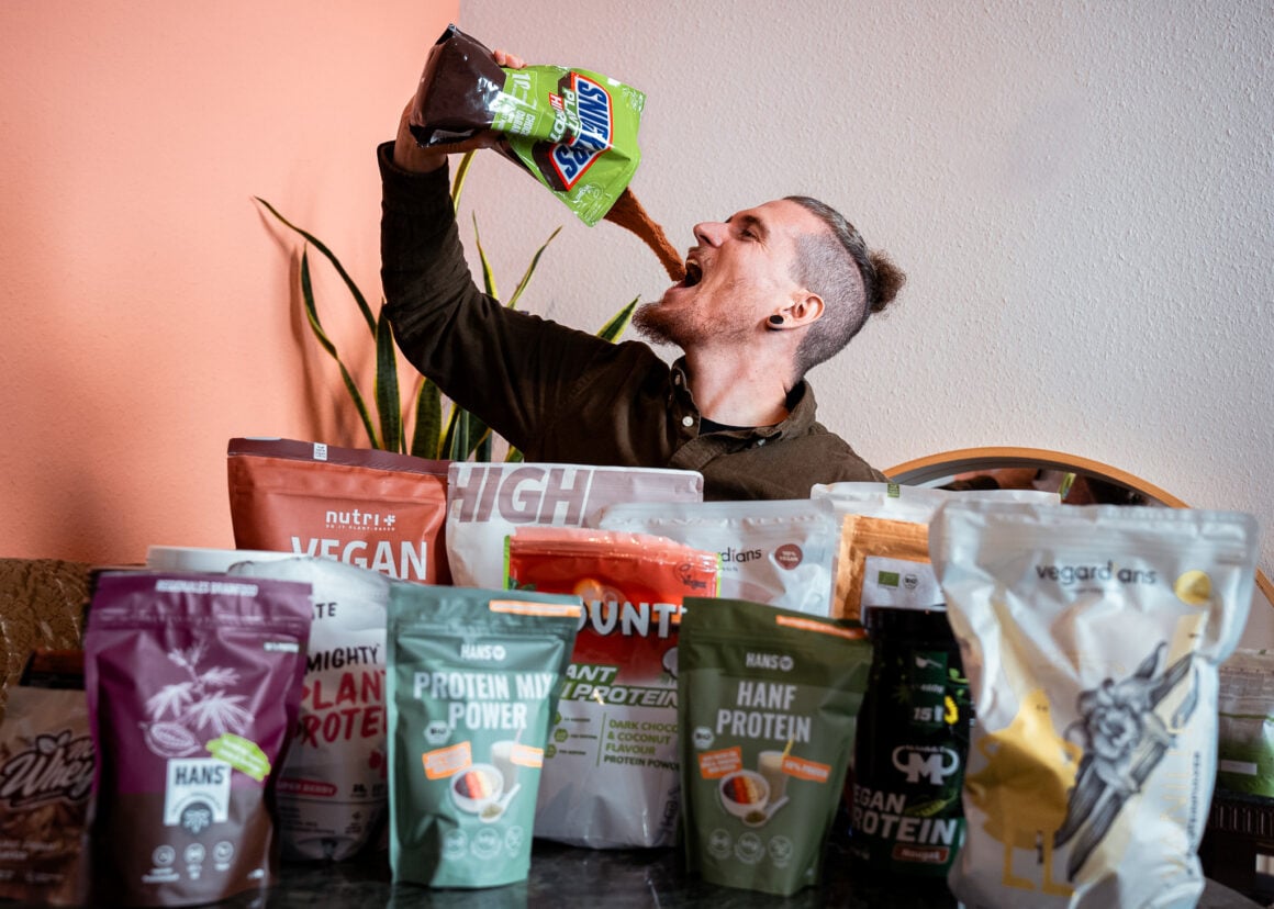Yannick (Gründer von This Is Vegan) sitzt hinter eine Menge an Verpackungen mit veganem Proteinpulver und hält eine Verpackung nach oben kippt sich Inhalt in den Mund