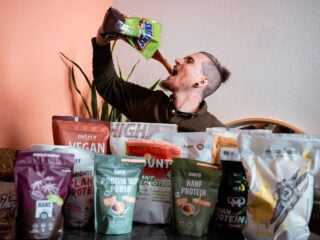Yannick (Gründer von This Is Vegan) sitzt hinter eine Menge an Verpackungen mit veganem Proteinpulver und hält eine Verpackung nach oben kippt sich Inhalt in den Mund