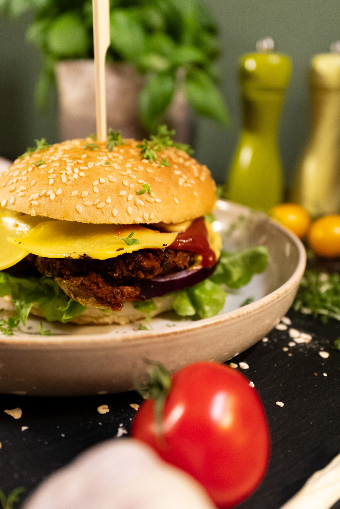 Teller auf dem ein veganer Burger mit veganem Käse zu sehen ist. Dahinter stehen Kräuter und es liegen Tomaten auf der Arbeitsplatte