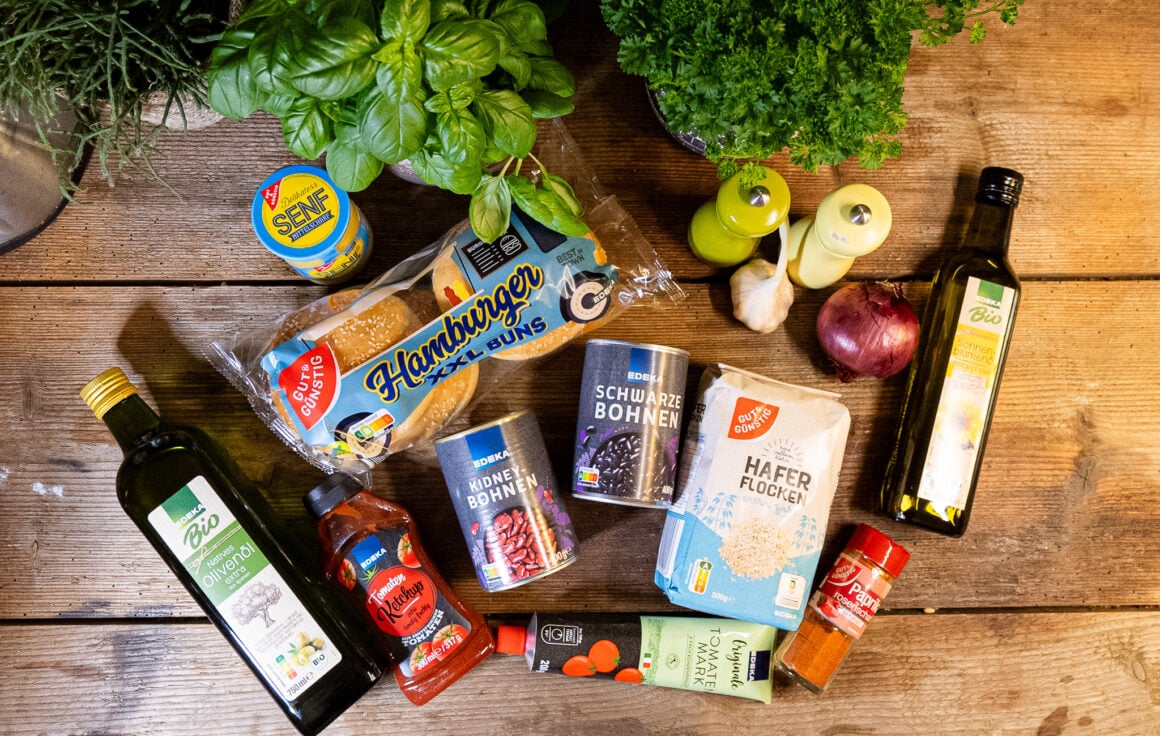 Lebensmittel wie Senf, Hamburger Brötchen, Kidneybohnen, schwarze Bohnen, Ketchup. Olivenöl, Tomatenmark, Haferflocken, Sonnenblumenöl, Paprikapulver, Knoblauch, Zwiebel, Salz und Pfeffer liegen auf einer Arbeitsplatte